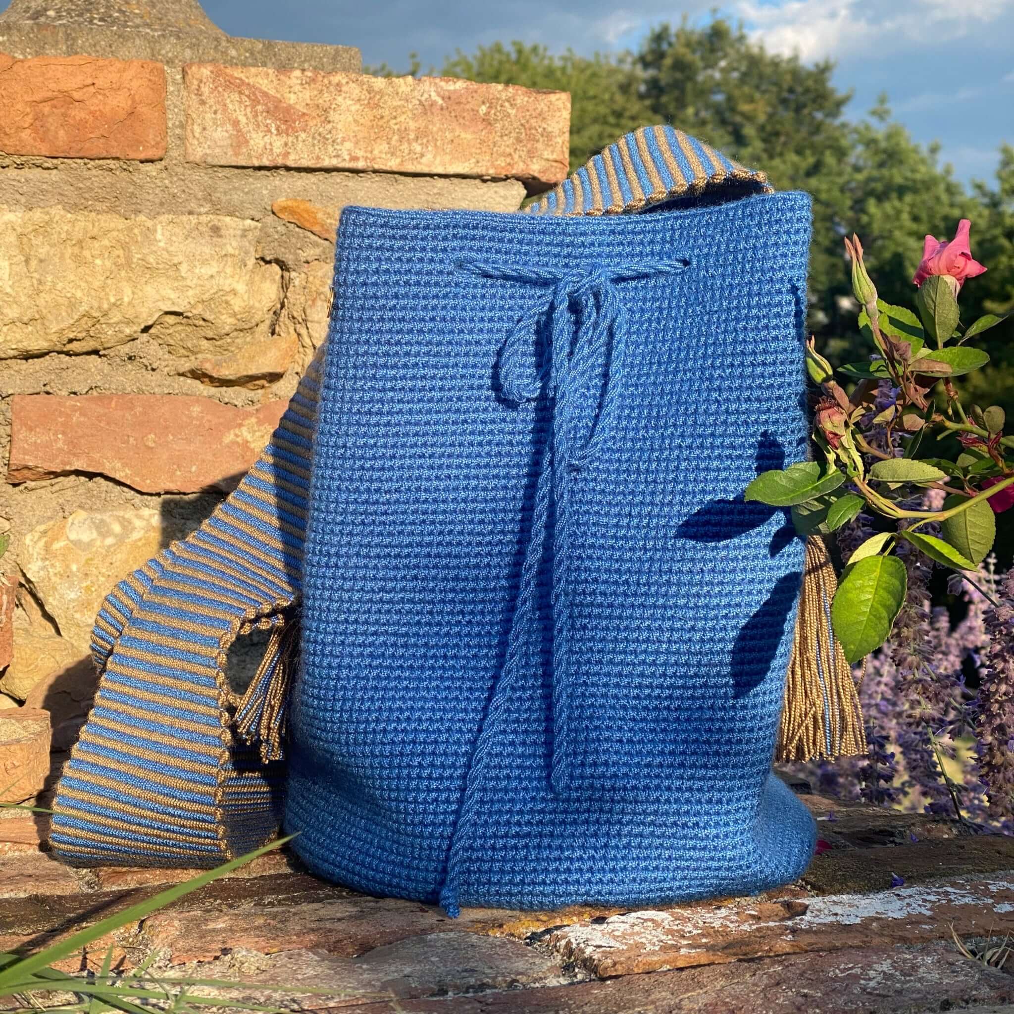 primo piano della borsa artigianale appoggiata ad un muretto con rose e lavanda in sottofondo. foto che mette in evidenza la lavorazione della mochila con trama intricata e tracolla in gro colbalto e grigio