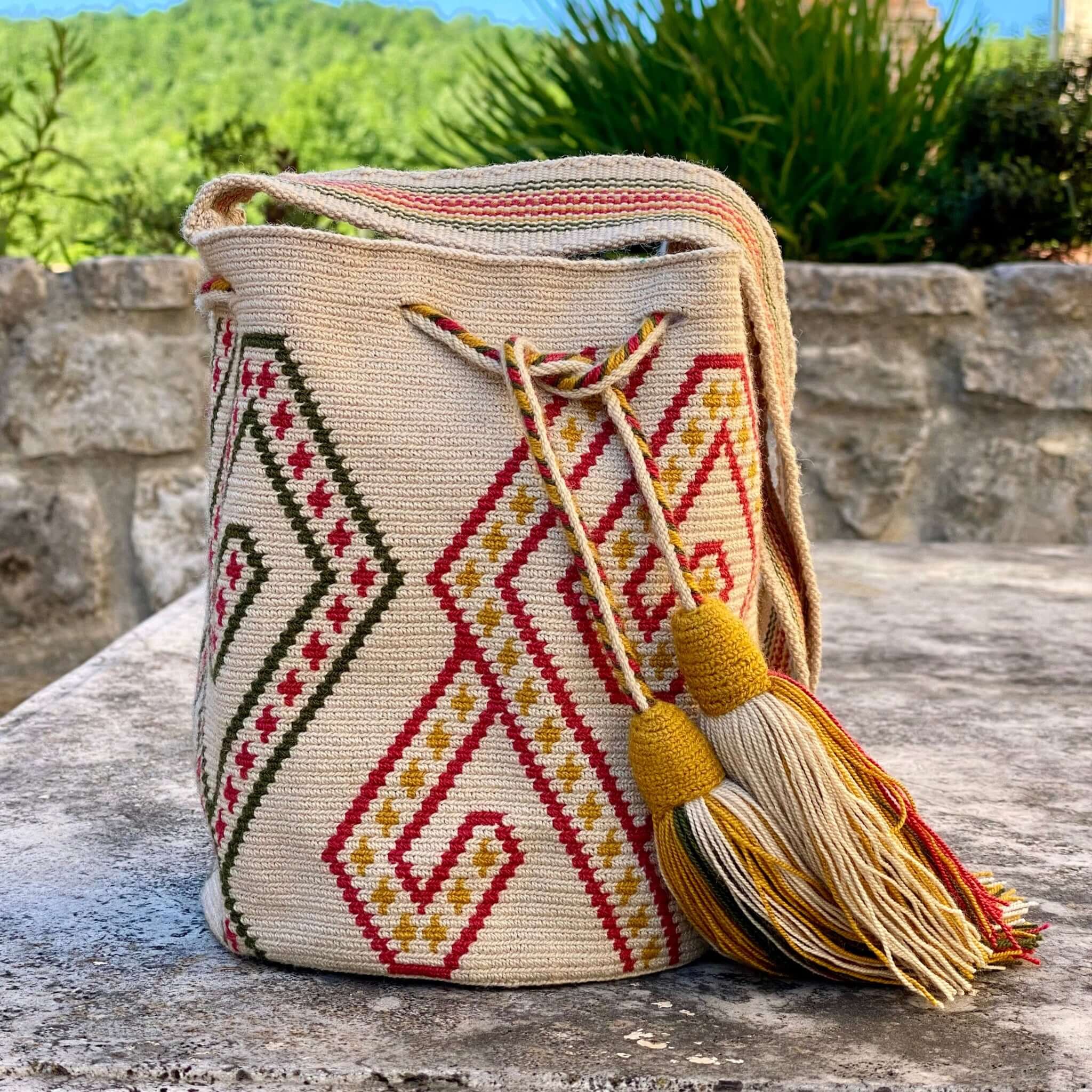 piccola borsa artigianale colombiana lavorata all'uncinetto con motivi tradizionali e schema geometrico. La borsa a secchiello con coulisse per chiuderla completamente e le nappine donano grazia alla praticità. la base è neutra color sabbia si sposa con le geometrie alternate in borgogna e verdone al cui interno sono ricamati dei piccoli rombi color ocra, verdone e bordeaux.