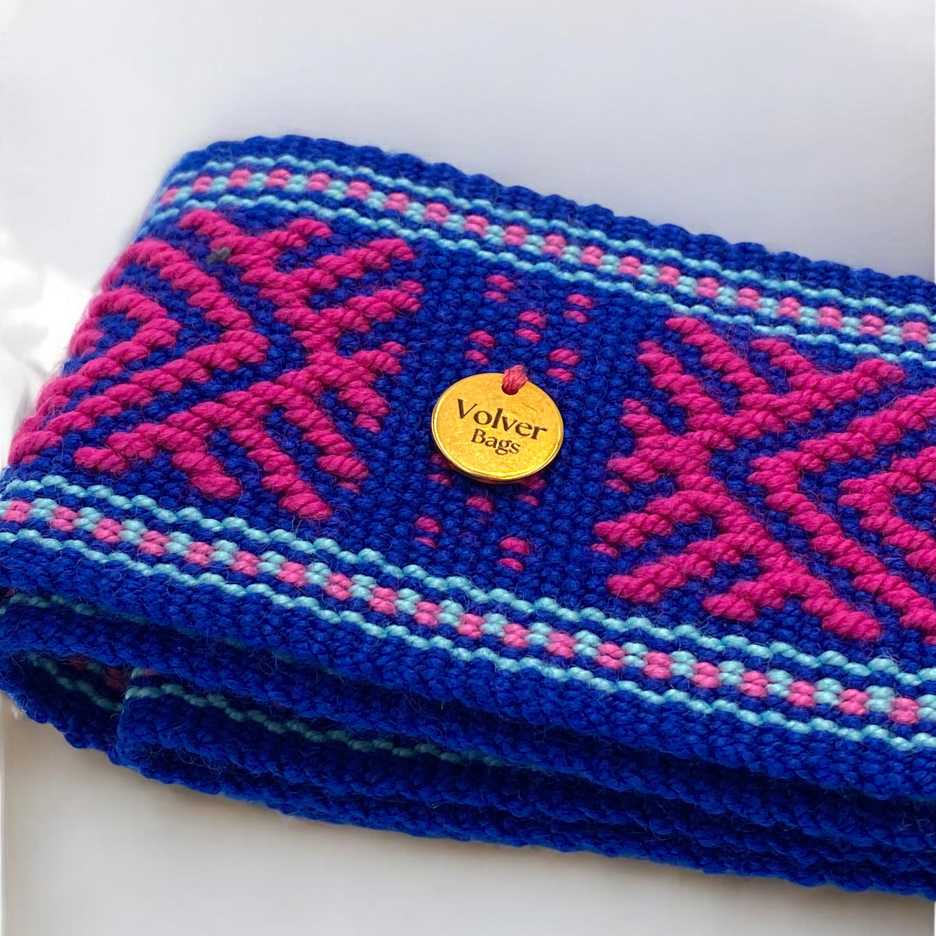 Macro dettaglio con medaglietta volver bags e ricamo della cintura artigianale tessuta a mano in cotone, colori blu, rosa e azzurro, realizzata da artigiane colombiane