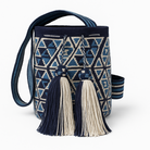 Borsa Wayuu blu e avorio con motivi geometrici e due grandi nappine, appoggiata su un tavolo rotondo di pietra in un giardino fiorito all’inglese, con sfondo verde e fiori colorati leggermente sfocati – Volver Bags.
