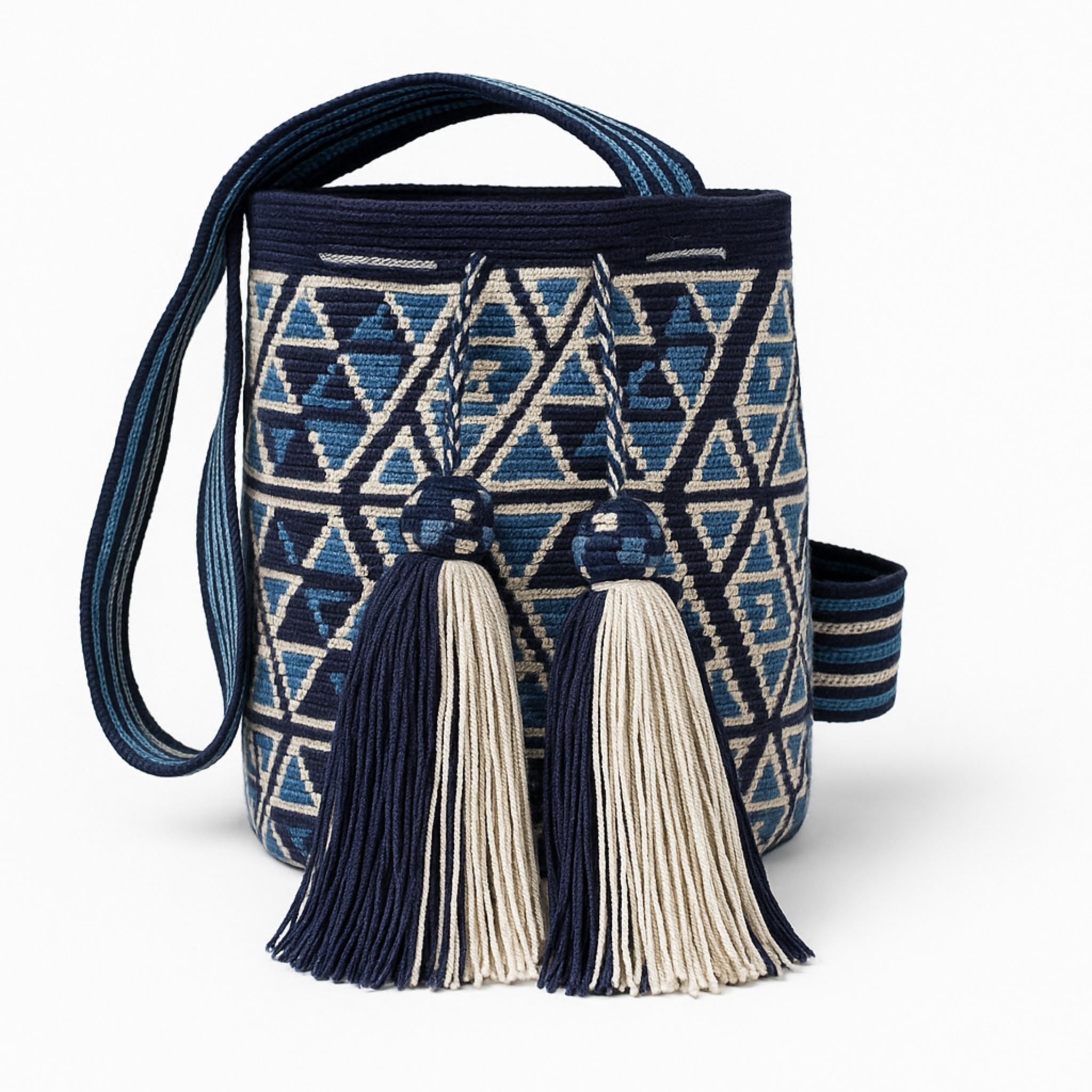 Borsa Wayuu blu e avorio con motivi geometrici e due grandi nappine, appoggiata su un tavolo rotondo di pietra in un giardino fiorito all’inglese, con sfondo verde e fiori colorati leggermente sfocati – Volver Bags.
