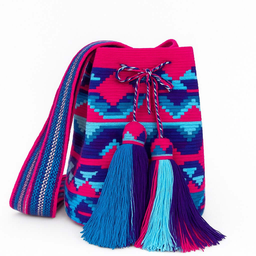 Borsa Wayuu intrecciata a filo singolo con motivi fucsia e azzurri, nappine rifinite e tracolla tessuta a mano – simbolo dell’artigianato autentico colombiano

